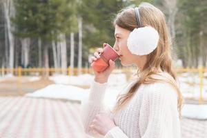 ung flicka dricker kaffe i en park. tonåring bär hörselkåpor och håller kaffe att gå. ta bort dryck koncept. sidovy foto