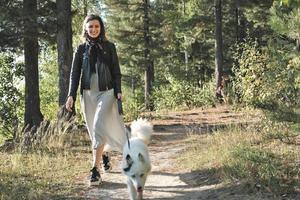 ung kvinna går i skogen med en hund. solig höstdag. leende kvinna klädd i klänning och jacka tittar på kameran och har kul med vit yakutisk laika medan du går längs passet foto