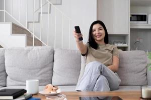 asiatisk ung vacker kvinna tittar på rolig film på tv hemma. attraktiv avslappnad tjej känner sig glad och slappna av, sitta på soffan ha roligt titta på komedivideo på tv i huset. aktivitet livsstilar koncept foto