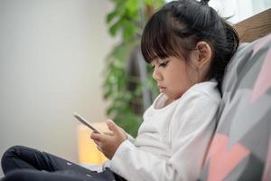 söt liten unge flicka tittar på video på smartphone med smiley ensam på sängen, barn som använder mobiltelefon med glad ansikte hemma. stanna hemma i karantän coronavirus covid-19 pandemi förebyggande. foto