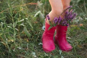 röda gummistövlar på fötterna av en flicka med en bukett vilda blommor. blommor i en känga, sommartid. sommar, frihet, natur, landsbygd, grönt gräs på fältet foto
