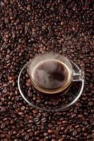 glas varmt svart kaffe med glasfat plats på högen av kaffefrö. varmt mörkt kaffe och ånga. närbild. toppvy. foto