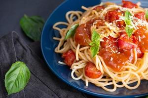 välsmakande aptitretande klassisk italiensk spaghettipasta med tomatsås, ostparmesan och basilika på tallrik och ingredienser för att laga pasta på mörkt bord. foto