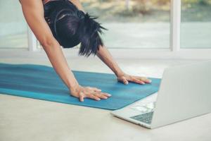 kvinna gör yoga nedåtvänd hund poserar inuti ljust mysigt rum genom fönster sett gröna lövträd sommar. stretching tittar på fitness video handledning online på laptop, tränar hemma sitter foto