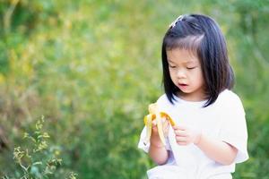 söt 4-årig asiatisk tjej äter banan till frukost. barn håller gul frukt i handen. bananskal barn. naturlig mat. barn bär vit t-shirt. tomt utrymme för att skriva in text. foto
