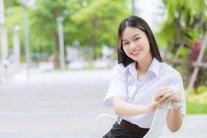 porträtt av vuxen thai student i universitetsstudentuniform. asiatisk vacker flicka sitter och ler glatt på utomhusuniversitet med en bakgrund av utomhusträdgårdsträd. foto