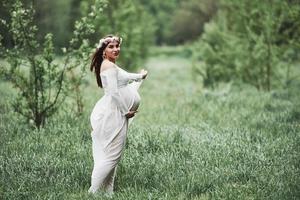 känna njutning. vacker gravid kvinna i klänning har en promenad utomhus. positiv brunett foto