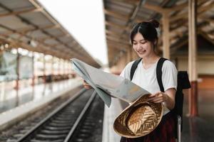 ung kvinna resenär med ryggsäck som vill kartlägga medan du väntar på tåget, asiatisk backpacker på järnvägsplattformen vid tågstationen. semester, resa, resa och sommar sommar resor koncept foto