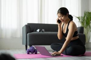 asiatisk sportig kvinna i sportkläder tränar och använder laptop och ringer telefon hemma i vardagsrummet, sitter på golvet med hantlar på yogamattan. sport och online träning koncept foto