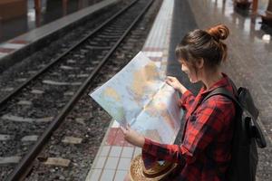 ung kvinna resenär med ryggsäck som vill kartlägga medan du väntar på tåget, asiatisk backpacker på järnvägsplattformen vid tågstationen. semester, resa, resa och sommar sommar resor koncept foto