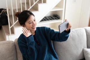 ung leende asiatisk kvinna sitta på mysig soffa håller mobiltelefon våg hand hälsning familj ringa videosamtal talar via virtuellt möte hemma. distanskommunikation vid karantän koncept foto