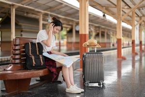 ung asiatisk kvinna resenär som sitter med karta väljer vart du ska resa och väska väntar på tåg på tågstationen, sommarsemester resekoncept foto