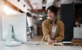 leende asiatisk ung kvinna använder headset tittar på laptop skärm lyssna och lära sig onlinekurser. glad affärskvinna med hörlurar videosamtal för kundtjänst foto