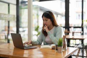 ung asiatisk kvinna bär hörlurar studera online titta på webbseminarium podcast på bärbar dator lyssna lärande utbildning kurs konferenssamtal göra anteckningar sitta vid skrivbordet, e-lärande koncept foto