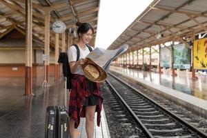 ung kvinna resenär med ryggsäck som vill kartlägga medan du väntar på tåget, asiatisk backpacker på järnvägsplattformen vid tågstationen. semester, resa, resa och sommar sommar resor koncept foto