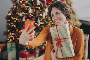 glad italiensk kvinna sitter nära vacker julgran hemma och tar selfie med present foto