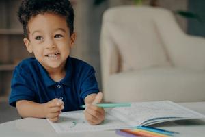 söt afroamerikansk liten pojke i mörkblå pikétröja målarbok hemma foto