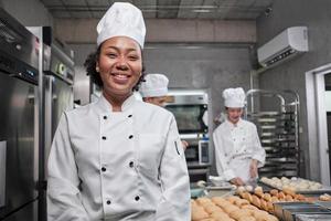 porträtt av ung afrikansk amerikansk kvinnlig kock i vit matlagningsuniform tittar på kameran, glada leende med mat professionell sysselsättning, kommersiella bakverk kulinariska jobb i ett restaurangkök. foto