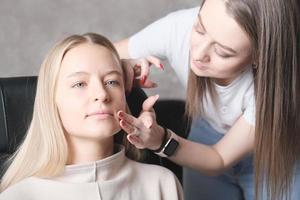 visagiste eller make-up artist använder fingrarna för att applicera cc cream på den unga kvinnans ansikte. blond vacker kvinna i skönhetsstudio foto