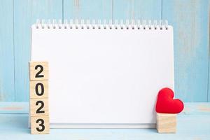 tom anteckningsbok och 2023 kuber med röd hjärtform dekoration på blå träbordsbakgrund. nytt år nytt du, mål, resolution, hälsa, kärlek och glad alla hjärtans dag-koncept foto