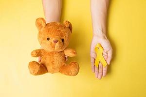 hand som håller gult band och björn docka på gul bakgrund för att stödja barn levande och sjukdom. september barndom cancer medvetenhet månad och världen cancer dag koncept foto