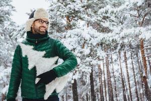 gladlynt trevlig man med skägg tittar eftertänksamt åt sidan, håller gran, bär glasögon, hatt och jacka, poserar mot vinterträd täckta med snö. människor och livsstilskoncept foto
