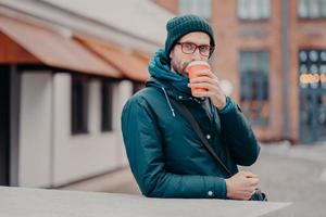 utomhusbild av stilig hipster dricker takeawy kaffe från engångsmugg, klädd i huvudbonader, jacka, bär glasögon, har srtoll utanför, tittar rakt på kameran. människor och livsstil foto