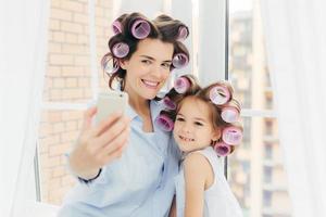 attraktiv liten unge med positivt uttryck, charmigt leende står nära sin mamma, gör selfie med modern mobiltelefon, posera mot fönsterbakgrund i rummet. föräldraskap och skönhetskoncept foto