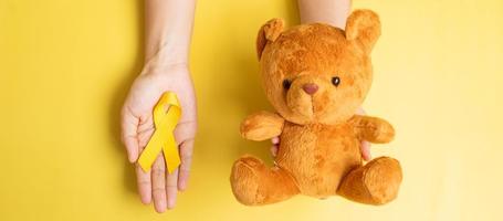 hand som håller gult band och björn docka på gul bakgrund för att stödja barn levande och sjukdom. september barndom cancer medvetenhet månad och världen cancer dag koncept foto