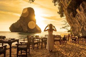 kvinna turist i vit klänning middag i restaurang grotta på phra nang stranden i solnedgången, railay, krabi, thailand. semester, resor, sommar, wanderlust och semester koncept foto