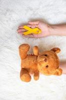 hand som håller gult band och björn docka på vit bakgrund för att stödja barn levande och sjukdom. september barndom cancer medvetenhet månad och världen cancer dag koncept foto