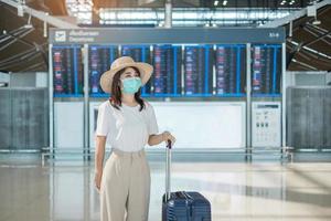 ung kvinna som bär ansiktsmask med bagage som går på flygplatsen, skydd mot coronavirus sjukdom, asiatisk kvinna resenär med hatt. tid att resa, testa och gå och vaccin boosterdos koncept foto