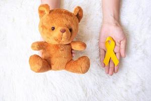 hand som håller gult band och björn docka på vit bakgrund för att stödja barn levande och sjukdom. september barndom cancer medvetenhet månad och världen cancer dag koncept foto