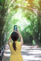 asiatisk kvinna i gul klänning och hatt som reser i grön bambutunnel, glad resenär som tar foto med mobiltelefon vid Chulabhorn wanaram-templet i Nakhon Nayok, Thailand