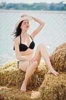 porträtt av en underbar tjej i svart bikinibaddräkt poserar på höbalen med en hatt vid sjön. foto