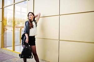 snygg brunett tjej slitage på skinnjacka och shorts med ryggsäck mot gyllene vägg. foto