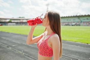 fitness sportig tjej i sportkläder på stadion utomhussporter. glad sexig kvinna dricksvatten från sport flaska mockup. foto