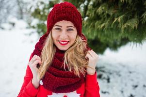 blond flicka i röd halsduk, hatt och jultomtens tröja poserar i parken på vinterdagen. foto