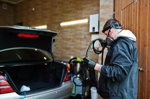 man i uniform och respirator, arbetare på biltvättcenter, städar bilinredning med varm ångtvätt. bil detaljering koncept. foto