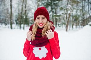 blond flicka i röd halsduk, hatt och jultomtens tröja poserar i parken på vinterdagen. foto