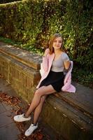 ung blond flicka i svart kjol och rosa kappa poserade på en solig dag på gatan. foto