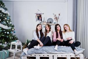 fyra söta vänner flickor bär på varma tröjor och svarta byxor på sängen på nyår inredda rum på studio. foto
