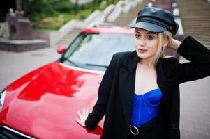 porträtt av vacker blond sexig modekvinna modell i mössa och i helsvart, blå korsett, med ljus smink nära röd stadsbil. foto
