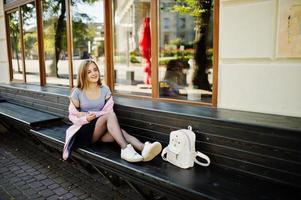 ung blond tjej i svart kjol och rosa kappa med kvinna ryggsäck sitter på bänken på staden. foto