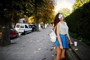 lockigt snygg tjej bär på blå jeans kjol, blus och glasögon poserade på gatan i staden med en kopp kaffe. foto