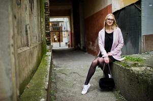 blond flicka vid glasögon och rosa kappa, svart tunika och handväska poserade på gatan. foto