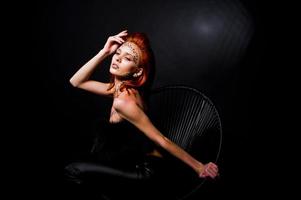modell rödhårig tjej med ursprungligen smink som leopard rovdjur isolerad på svart. studio porträtt på stol. foto