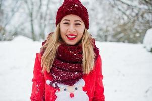 blond flicka i röd halsduk, hatt och jultomtens tröja poserar i parken på vinterdagen. foto