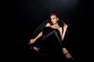modell rödhårig tjej med ursprungligen smink som leopard rovdjur isolerad på svart. studio porträtt på stol. foto
