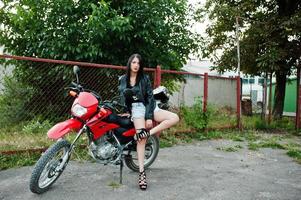 porträtt av en cool och fantastisk kvinna i klänning och svart skinnjacka som sitter på en cool röd motorcykel. foto